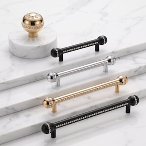 Könnte beinhalten: Eine Kollektion von Möbelbeschlägen, bestehend aus einem kugelförmigen goldenen Knopf und vier länglichen Stangengriffen in Schwarz, Silber und Gold. Jedes Teil verfügt über ein zentrales Band aus eingebetteten klaren Kristallen entlang der Metalloberfläche.