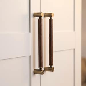 Puede incluir: Dos tiradores de armario verticales con una empuñadura cilíndrica de madera marrón oscuro centrada entre dos soportes de montaje de metal de latón cepillado. Los tiradores están fijados a una puerta blanca con paneles y diseño lineal.