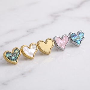 May include: Five heart-shaped pins arranged in a row. The first is gold-toned with iridescent green inlay, the second is gold-toned with white mother-of-pearl, the third is solid gold-toned, the fourth is silver-toned with pink, and the fifth is silver-toned with iridescent blue inlay.