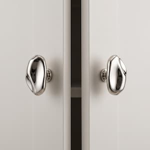 May include: Two polished silver cabinet knobs with an irregular, organic, pebble-like shape. Each knob features a smooth, reflective metallic surface and is mounted onto a white cabinet door, providing a distinct contrast to the flat white finish.