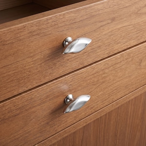May include: Two polished silver-toned metal drawer knobs with an elongated, smooth, and slightly curved leaf-like shape. These hardware pieces are mounted onto the front panels of a wooden cabinet featuring a visible light brown wood grain pattern.