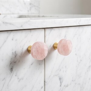 Puede incluir: Dos pomos redondos para armarios con caras de piedra de cuarzo rosa claro pulido fijadas a bases cil&iacute;ndricas de lat&oacute;n cepillado, montados en puertas de armario de m&aacute;rmol blanco con vetas grises en toda la superficie.