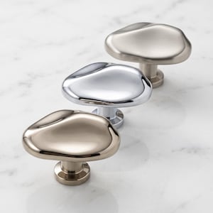 K&ouml;nnte beinhalten: Drei Schrankkn&ouml;pfe mit unregelm&auml;&szlig;igen, organischen, kieselsteinartigen Formen. Der vordere Knopf hat ein poliertes Champagner-Finish, der mittlere Knopf ist aus reflektierendem Chrom und der hintere Knopf hat ein geb&uuml;rstetes Nickel-Finish.