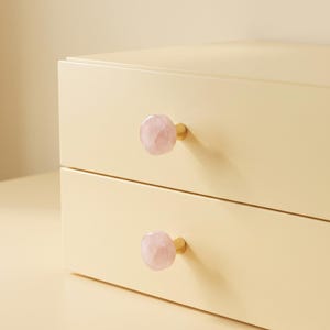 Puede incluir: Una unidad de almacenamiento de color crema claro con dos cajones horizontales, cada uno con un pomo de piedra rosa p&aacute;lido facetado unido a una peque&ntilde;a base de metal dorado. Los cajones son lisos y rectangulares con bordes rectos.