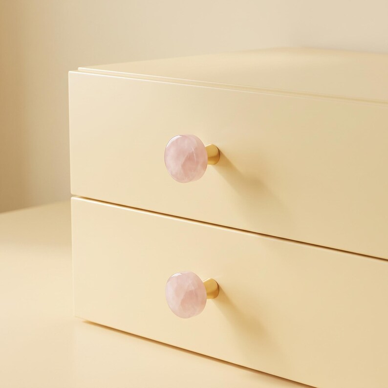 Puede incluir: Una unidad de almacenamiento de color crema claro con dos cajones horizontales, cada uno con un pomo de piedra rosa p&aacute;lido facetado unido a una peque&ntilde;a base de metal dorado. Los cajones son lisos y rectangulares con bordes rectos.