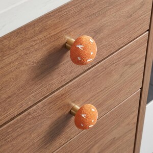 May include: Two round cabinet knobs with a textured orange surface and white flecks, mounted on a wooden drawer front. Each knob features a gold-toned metal base that connects the spherical handle to the wood surface of the furniture piece.
