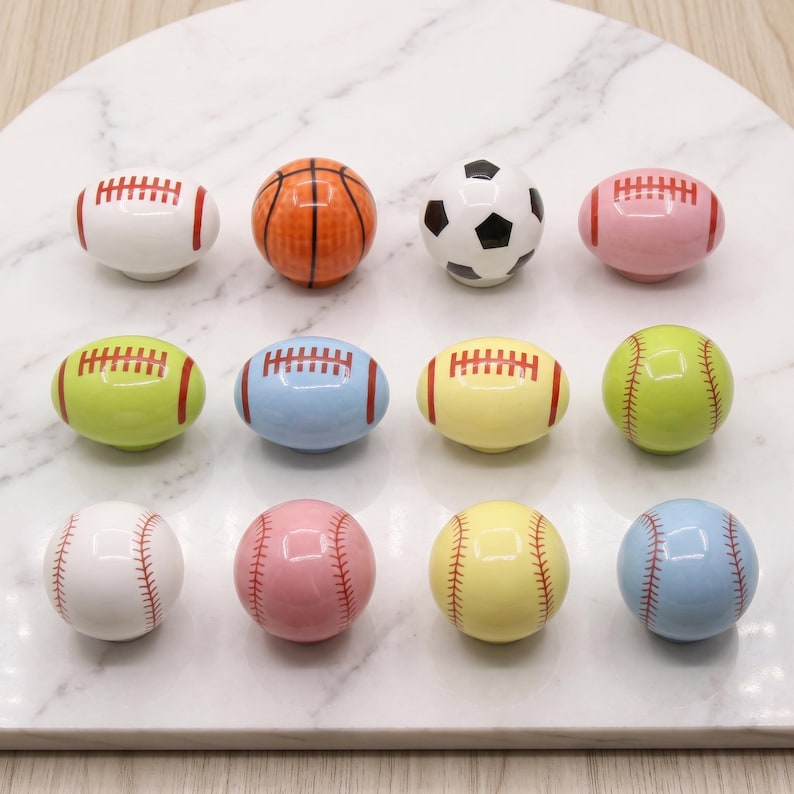 May include: A collection of twelve ceramic cabinet knobs shaped like various sports balls, including footballs, basketballs, soccer balls, and baseballs in assorted colors such as white, orange, pink, lime green, yellow, and light blue.
