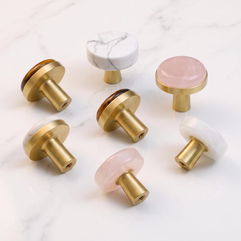 Puede incluir: Un conjunto de siete pomos circulares para armarios con bases de metal dorado. Los pomos tienen superficies superiores variadas, incluyendo m&aacute;rmol blanco con vetas grises, blanco s&oacute;lido, rosa p&aacute;lido transl&uacute;cido y patrones de piedra ojo de tigre.