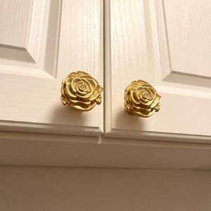 May include: Two gold-colored cabinet knobs shaped like blooming roses. Each knob features detailed, layered petals and a rounded form, mounted onto the surface of white, paneled cabinet doors with a visible wood grain texture.