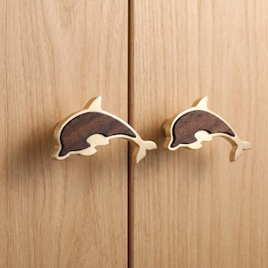 May include: Two cabinet knobs shaped like dolphins, each featuring a dark brown wood inlay surrounded by a polished gold-tone metal frame. The dolphin-shaped handles are mounted horizontally on a light-coloured wood cabinet door surface.