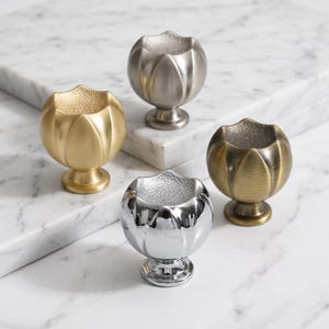 May include: Four metal cabinet knobs shaped like flower buds with a textured interior. The set includes one brushed gold, one brushed silver, one polished chrome, and one antique brass finish, each featuring a rounded base and a stem.