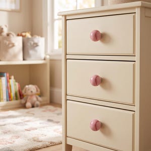 May include: A cream-colored wooden dresser with three drawers, each featuring a round, pink baseball-shaped knob. The knobs have a glossy finish and are detailed with red stitching patterns, providing a distinct contrast to the neutral furniture.