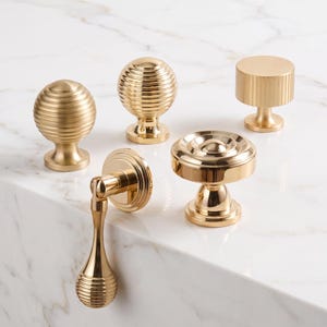 May include: A collection of five gold-toned cabinet hardware pieces, including four round knobs with various ribbed and smooth textures, and one drop-style pull with a matching ribbed finish on a circular base, all featuring a polished metallic sheen.