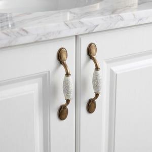 May include: Two cabinet pulls featuring an antique brass finish with a central white ceramic insert. The ceramic section displays a distinct black crackle pattern. These hardware pieces are mounted vertically on white cabinet doors with panels.