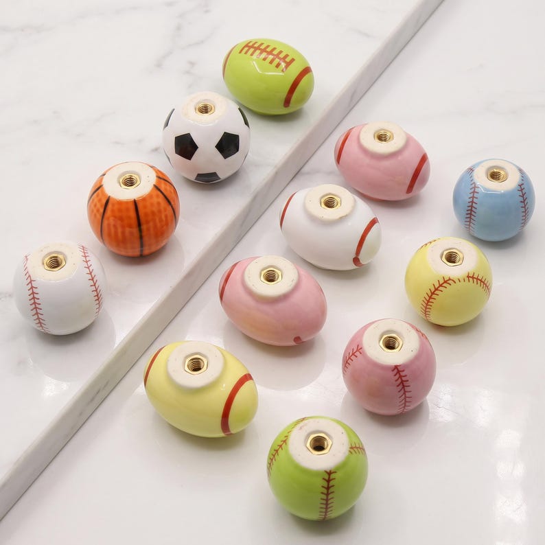 May include: A collection of eleven ceramic cabinet knobs shaped like various sports balls, including baseballs, basketballs, soccer balls, and footballs. Each knob features a central brass-colored metal threaded insert for mounting purposes.