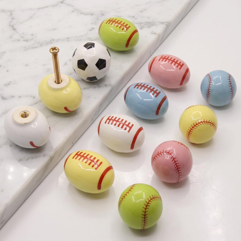 May include: A collection of ceramic cabinet knobs shaped like various sports balls, including footballs, soccer balls, and baseballs in colors like yellow, white, pink, blue, and green. Each knob features a metal screw for mounting purposes.