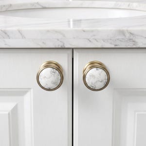 Pode incluir: Dois puxadores redondos para arm&aacute;rios com um padr&atilde;o de m&aacute;rmore branco e veios cinzentos, inseridos numa moldura circular de metal escovado em tom de lat&atilde;o. Estas pe&ccedil;as est&atilde;o montadas em portas de arm&aacute;rio brancas sob uma bancada de m&aacute;rmore.
