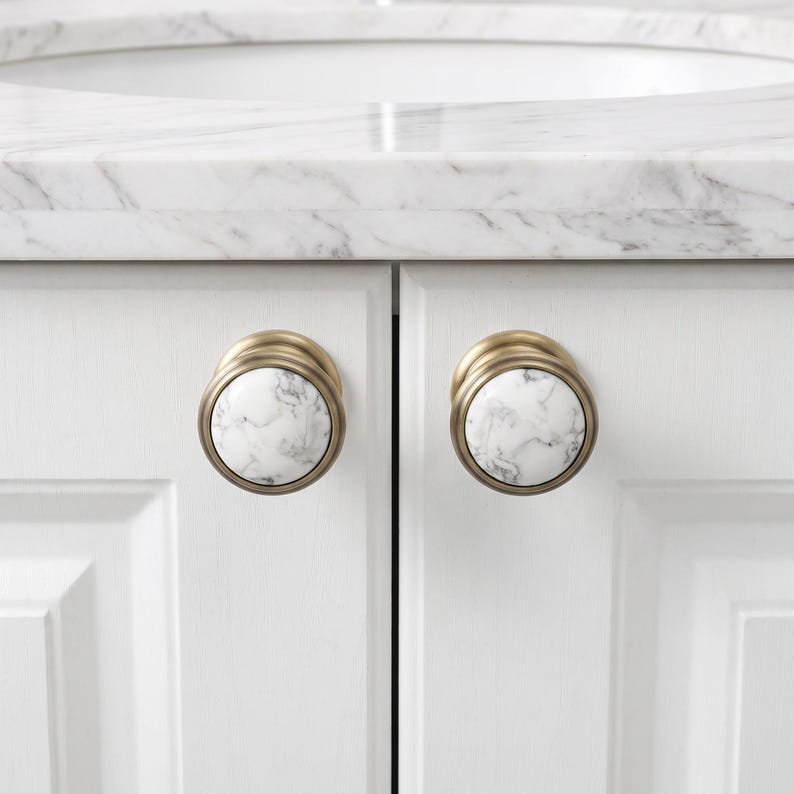 Pode incluir: Dois puxadores redondos para arm&aacute;rios com um padr&atilde;o de m&aacute;rmore branco e veios cinzentos, inseridos numa moldura circular de metal escovado em tom de lat&atilde;o. Estas pe&ccedil;as est&atilde;o montadas em portas de arm&aacute;rio brancas sob uma bancada de m&aacute;rmore.