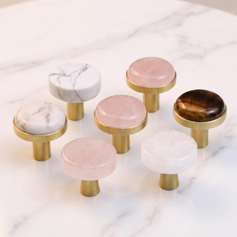 Puede incluir: Una colecci&oacute;n de siete pomos para armarios con tapas de piedra circulares en varios colores, incluyendo m&aacute;rmol blanco, rosa p&aacute;lido y marr&oacute;n oscuro, cada uno montado sobre una base cil&iacute;ndrica de lat&oacute;n cepillado sobre una superficie blanca.