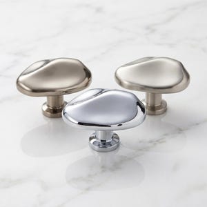 K&ouml;nnte beinhalten: Drei Schrankkn&ouml;pfe mit unregelm&auml;&szlig;igen, organischen, kieselsteinartigen Formen. Ein Knopf hat eine polierte Chromoberfl&auml;che, w&auml;hrend die anderen beiden geb&uuml;rstete Metalloberfl&auml;chen aufweisen. Jeder Knopf hat einen zylindrischen Sockel.