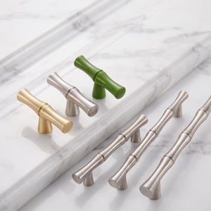 May include: A collection of cabinet hardware featuring T-shaped knobs and long pulls with a bamboo-inspired design. The set includes a brushed gold knob, a brushed silver knob, a green knob, and three brushed silver pulls of varying lengths.