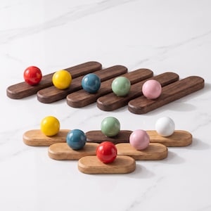 May include: A collection of ten wooden wall hooks, each featuring a smooth, spherical knob in a solid colour. Five hooks are made of dark walnut wood, and five are made of lighter oak wood. The knobs are red, yellow, blue, green, pink, and white.