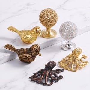 May include: A collection of decorative cabinet knobs and pulls featuring two bird-shaped knobs in gold and bronze, two spherical knobs with geometric lattice patterns in gold and silver, and two octopus-shaped knobs in gold and dark bronze finishes.