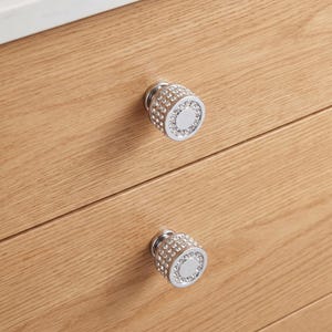 Op de afbeelding: Twee ronde kastknoppen met een gepolijste zilveren afwerking, voorzien van een gladde centrale schijf omringd door meerdere rijen kleine, heldere, gefacetteerde kristallen ingebed in het metalen oppervlak, gemonteerd op lichte houten lades.