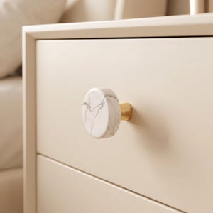 Puede incluir: Un pomo de mueble circular con una cara de m&aacute;rmol blanco con vetas grises, unido a una base cil&iacute;ndrica de metal dorado. El pomo est&aacute; montado en un frente de caj&oacute;n de madera de color beige claro, con un acabado liso y un dise&ntilde;o sencillo.