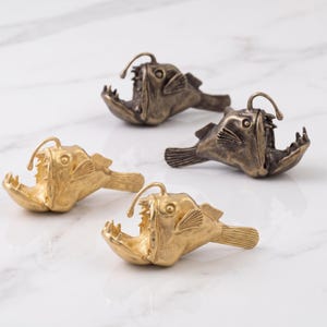 May include: Four anglerfish figurines arranged in a group. Two of the fish are finished in a bright gold tone, while the other two feature a darker, antique bronze patina. Each fish has an open mouth with sharp teeth and a prominent dorsal lure.