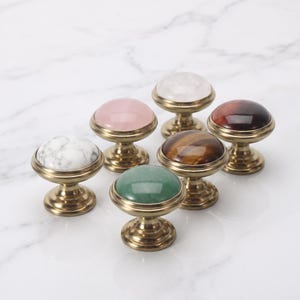 Pode incluir: Um conjunto de seis puxadores redondos para arm&aacute;rios com incrusta&ccedil;&otilde;es de pedra polida em m&aacute;rmore branco, rosa claro, transparente, castanho escuro, olho de tigre e verde, em bases met&aacute;licas circulares de lat&atilde;o envelhecido com pedestal.