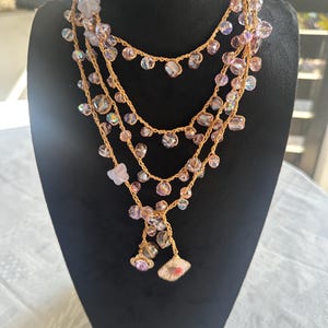 May include: A multi-strand necklace with gold-colored chain and iridescent beads. The necklace is displayed on a black mannequin. The beads are various sizes and shapes, with a mix of pink, purple, and clear colors. The necklace has a delicate, feminine design.