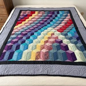 May include: A handmade quilt with a geometric design in a variety of colors, including red, yellow, blue, and purple. The quilt has a black border and a gray patterned edge. The quilt is displayed on a bed.