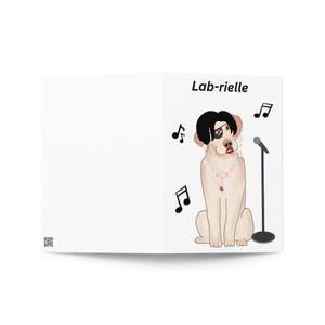 May include: A white greeting card featuring a cartoon illustration of a light brown Labrador dog wearing a black wig, an eye patch, pearl earrings, and a pearl necklace with a pink pendant. The text Lab-rielle appears above the dog.