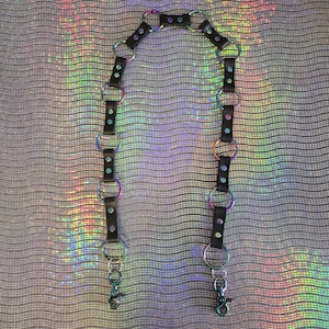 May include: Black leather harness with iridescent metal rings and hardware. The harness features a series of connected rings and straps, with lobster clasps at the ends. The metal rings have a rainbow sheen, and the background is a shimmering, iridescent fabric.