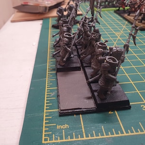 以下が含まれることがあります： ダークグレーで塗装されたファンタジー軍のミニチュアフィギュアが、黒いベースに配置されています。 フィギュアは槍と鎧を装備し、センチメートル単位の測定値を持つ緑色のカッティングマットに配置されています。