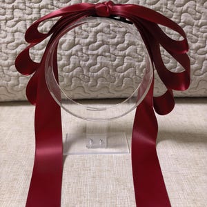 Op de afbeelding: Een haaraccessoire van donkerrood satijnen lint met een grote strik met meerdere lussen aan de bovenkant. De gladde, glanzende stof vormt aan beide kanten verschillende trapsgewijze lussen, met twee lange, rechte linten die naar beneden hangen.