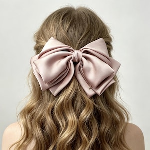 Può includere: Un grande fiocco per capelli color rosa polvere, realizzato in un tessuto liscio simile al raso. Il fiocco presenta molteplici anelli sovrapposti e un dettaglio a nodo centrale, fissato su capelli ondulati castano chiaro. Un piccolo ciondolo pende.