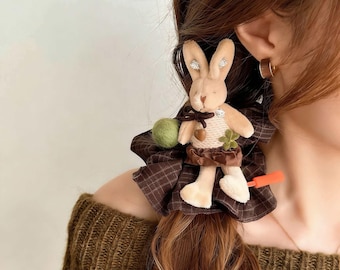 Puede incluir: Accesorio para el cabello de conejito de peluche marrón y beige con falda a cuadros, sosteniendo una bola verde y una zanahoria. El conejito tiene un lazo marrón y un trébol. El accesorio para el cabello está unido a una tela a cuadros marrón.