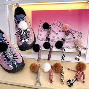May include: Pink suede trainers with black soles and blue detailing, decorated with pom-poms and small doll-like figures. A pink backdrop shows a shoe outline, and several pom-pom keychains are displayed.