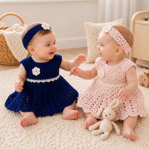 May include: Two infants wearing crocheted dresses and matching headbands. One infant wears a navy blue dress with a white flower accent and a navy headband. The other wears a pale pink dress with a white flower accent and a pink headband.