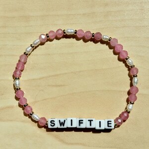 May include: A beaded bracelet with the word "SWIFTIE" spelled out in white letter beads. The bracelet features pink faceted beads, small white pearls, and gold-colored spacers. The bracelet is on a light wood surface.