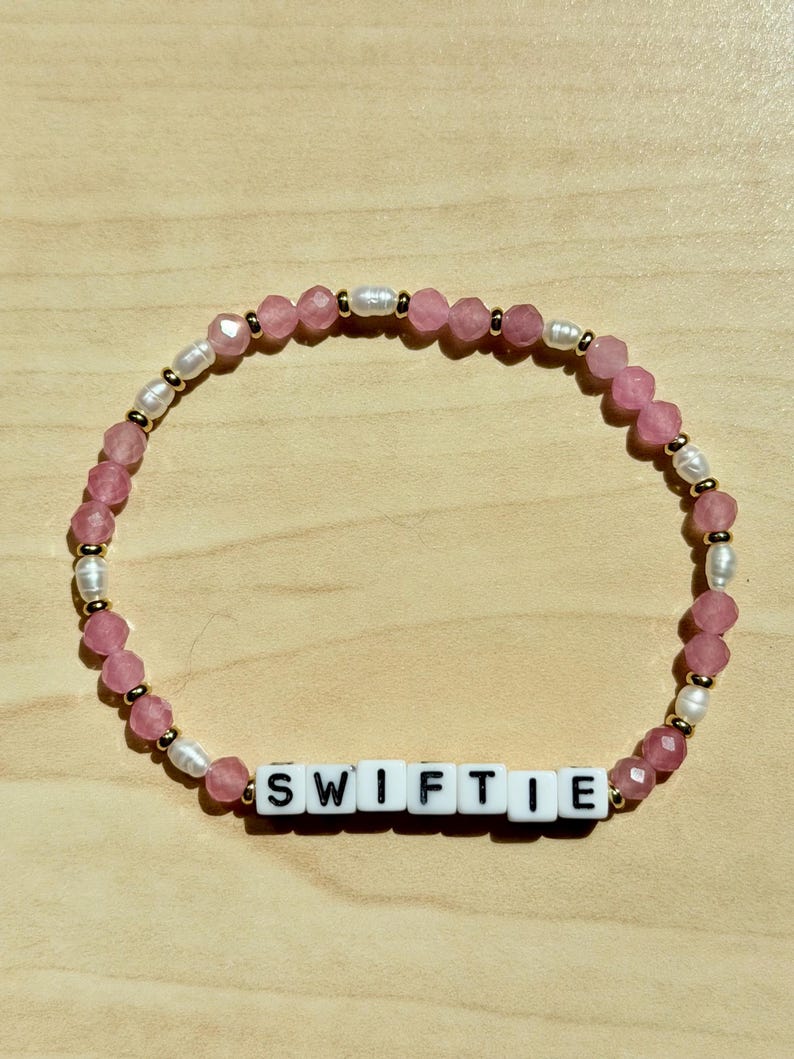 May include: A beaded bracelet with the word "SWIFTIE" spelled out in white letter beads. The bracelet features pink faceted beads, small white pearls, and gold-colored spacers. The bracelet is on a light wood surface.