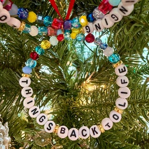 May include: A circular ornament made of multicolored translucent beads and white letter beads spelling out the phrase BRINGING THINGS BACK TO LIFE, accented with small gold-toned spacers and suspended by a thin red ribbon loop.