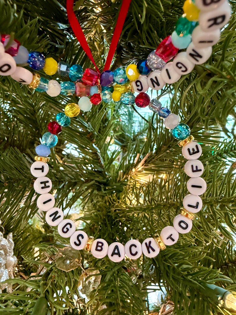May include: A circular ornament made of multicolored translucent beads and white letter beads spelling out the phrase BRINGING THINGS BACK TO LIFE, accented with small gold-toned spacers and suspended by a thin red ribbon loop.