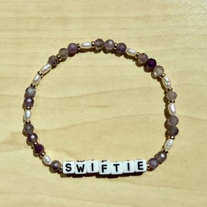 May include: A beaded bracelet with the word "SWIFTIE" spelled out in white letter beads. The bracelet features a mix of purple, white, and gold beads. The beads are arranged in a circular pattern, creating a stylish accessory.