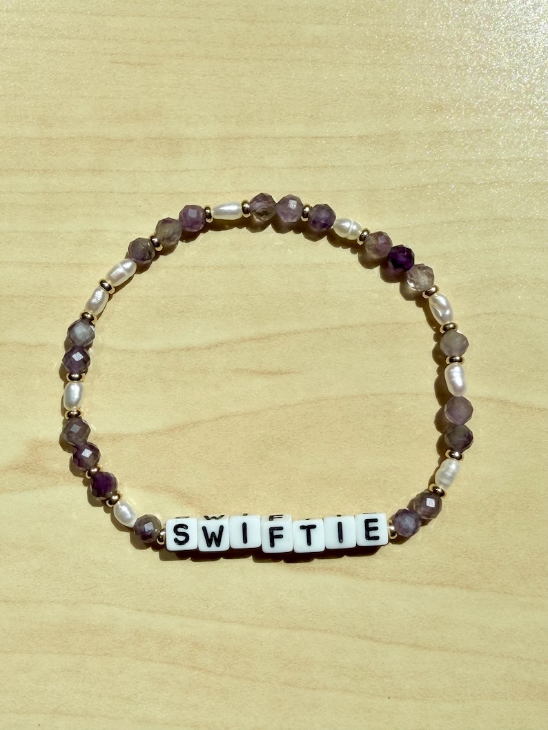 May include: A beaded bracelet with the word "SWIFTIE" spelled out in white letter beads. The bracelet features a mix of purple, white, and gold beads. The beads are arranged in a circular pattern, creating a stylish accessory.