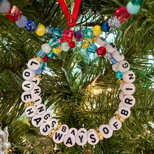 May include: A circular ornament made of white letter beads spelling LOVE HAS WAYS OF BRINGING THINGS, interspersed with gold-tone spacers and colorful translucent beads. A red ribbon loop is attached to the top for hanging on a tree.