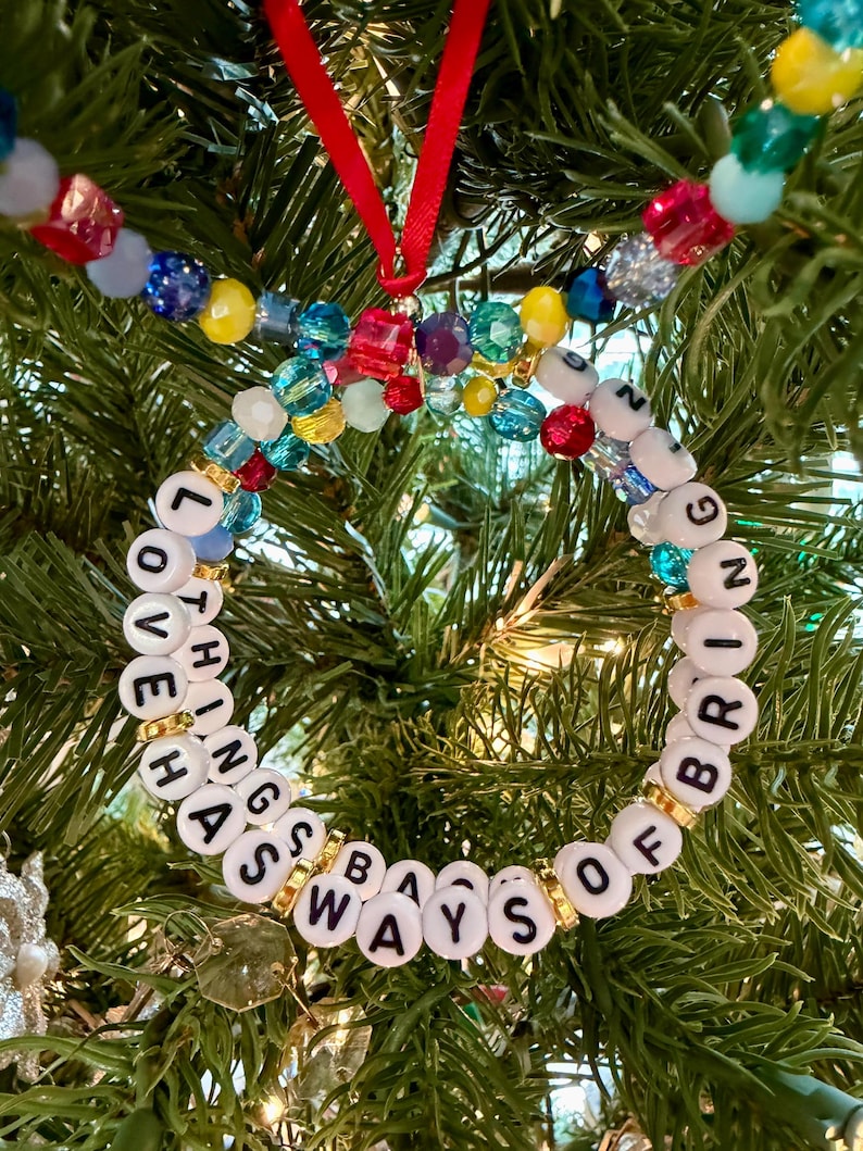 May include: A circular ornament made of white letter beads spelling LOVE HAS WAYS OF BRINGING THINGS, interspersed with gold-tone spacers and colorful translucent beads. A red ribbon loop is attached to the top for hanging on a tree.