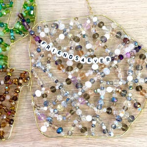 May include: Decorative beaded ornaments in the shape of a leaf and a geometric shape. The geometric shape has the text "FRIENDS4EVER" spelled out in white letter beads. The ornaments are made with gold wire and various colored beads.